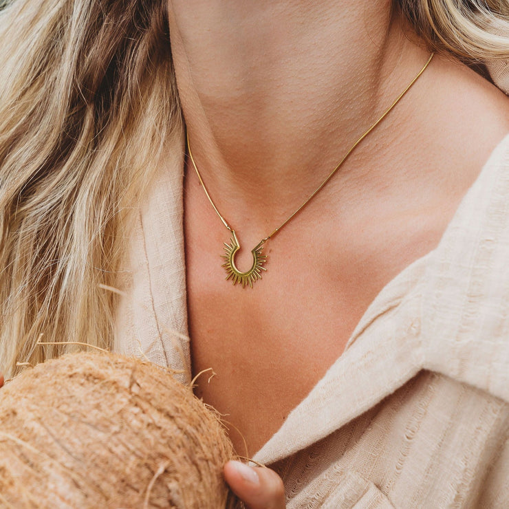 Blonde smiling model holding a coconut with Dainty gold necklace chain with a medium sized circular flat sun pendant and small white hugger hoops.