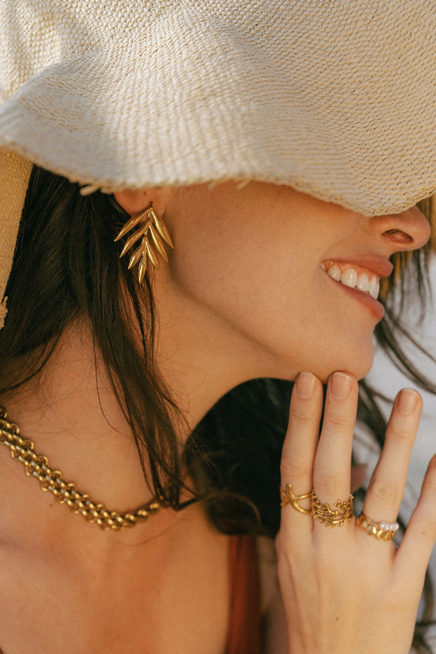 girl wearing tan hat, hand on her chin with gold rings on, a thick gold chain necklace and large gold leaf earrings