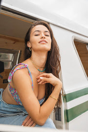 Brunette model with a van backdrop wearing a gold bracelet, gold hoop earrings, and a dainty gold choker made up of small flower pieces connected to make a necklace.
