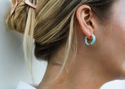 Product focused photo of a blonde model wearing small circular hugger hoops in aqua blue coloring with gold features.