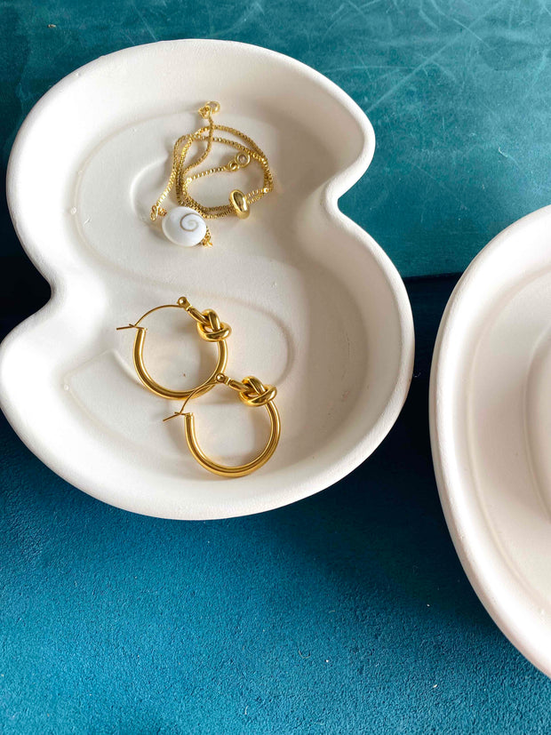 Gold love knot earrings with see shell bracelet in a white dish.